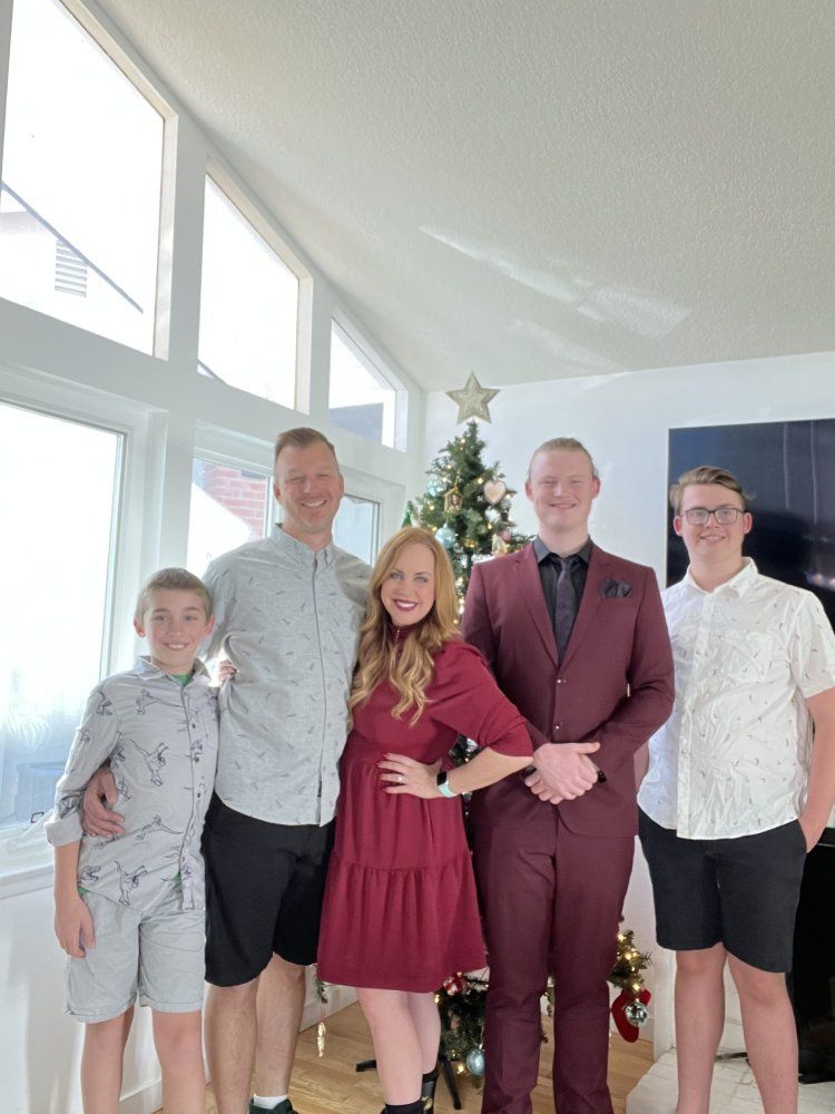 Family posing together by a Christmas tree; indoors with large windows. The woman is wearing a red dress.