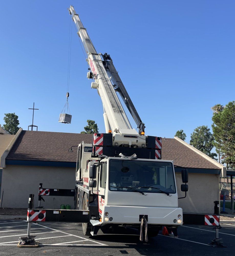 Crane lifting a gray object near a building with a brown roof under a blue sky.