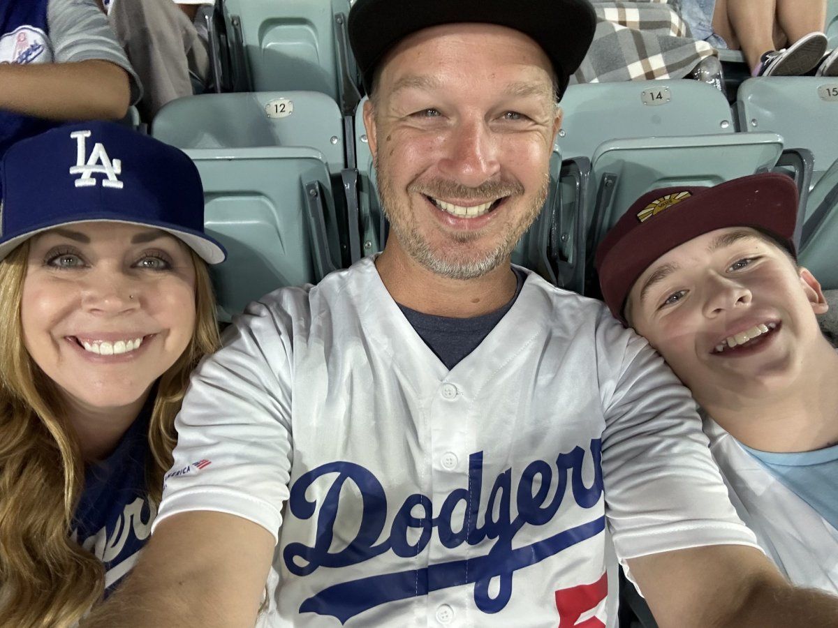 Family smiling at a Dodgers game: woman in cap, man in jersey, teen in cap.
