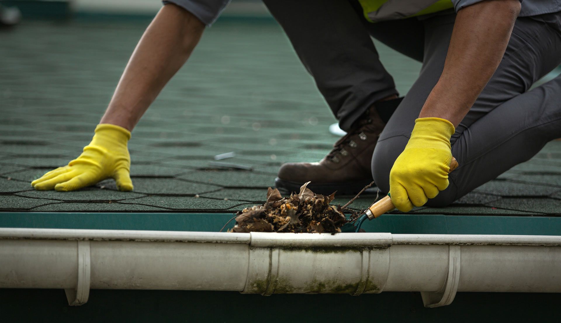 A man wearing yellow gloves is cleaning a gutter on a roof