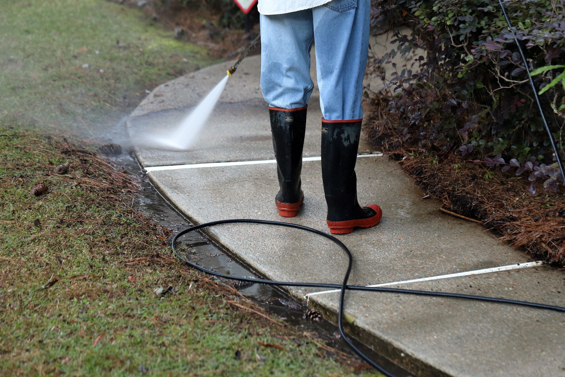 A person in jeans and black rubber boots pressure washes a concrete walkway next to a lawn and garden bed.