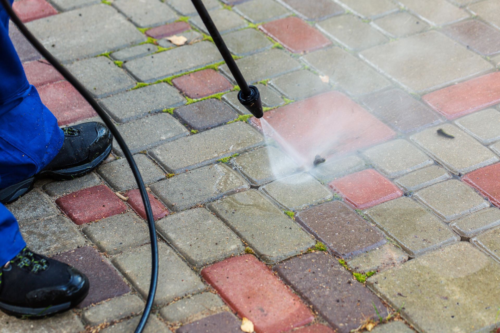 A person in blue work pants uses a pressure washer to clean a dirty paved walkway with red and grey bricks.