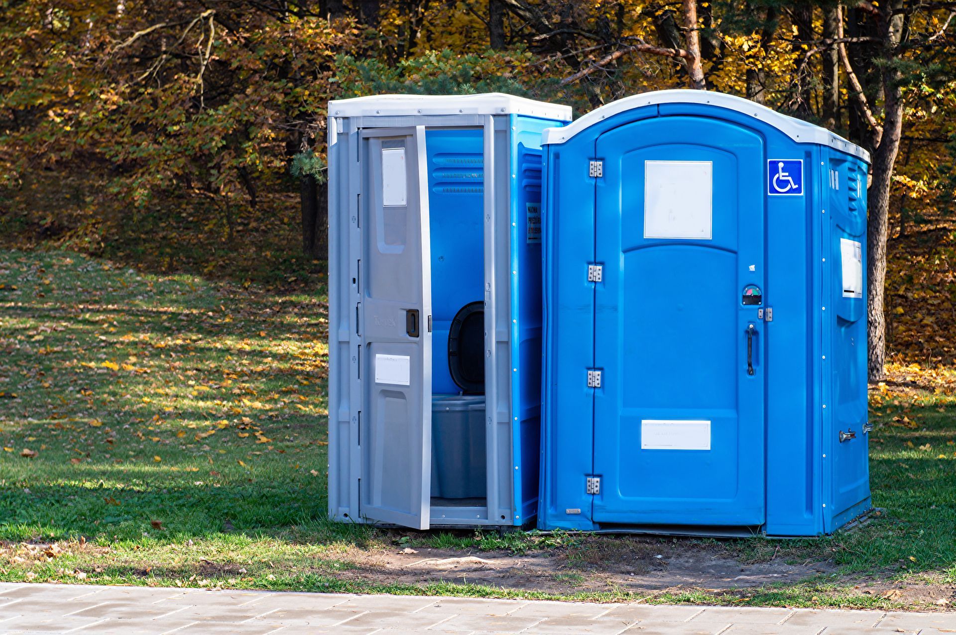 Two blue portable toilets standing on grass near a paved path, with one door open.