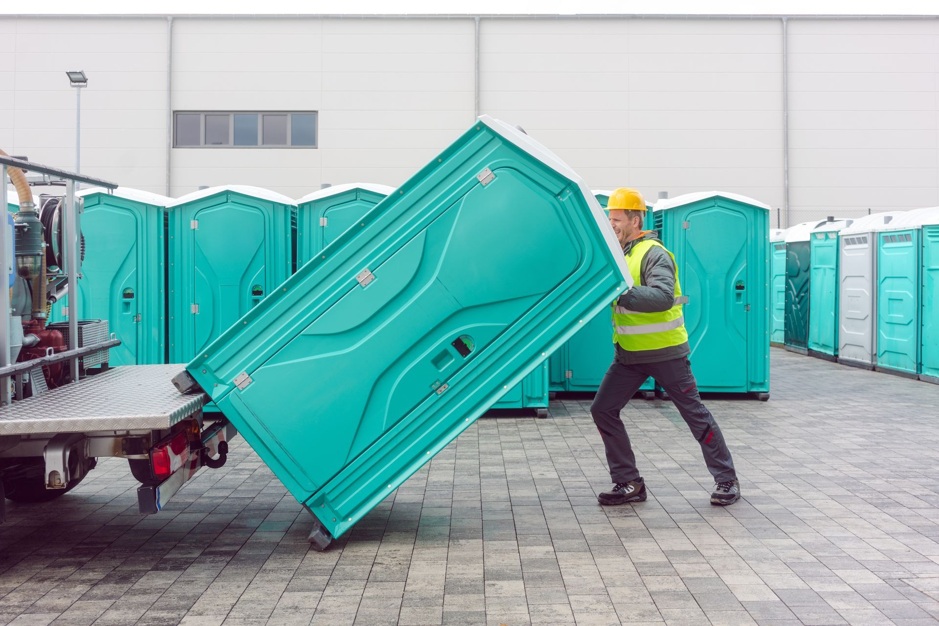 Worker unloading a portable toilet from a truck, with multiple portable restrooms in the background. Worker unloading a portable toilet from a truck, with multiple portable restrooms in the background.