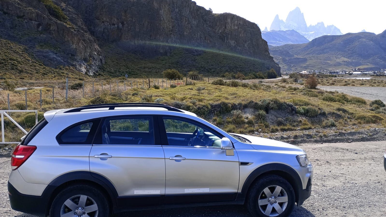 Un coche plateado está aparcado en una zona de grava con montañas al fondo.