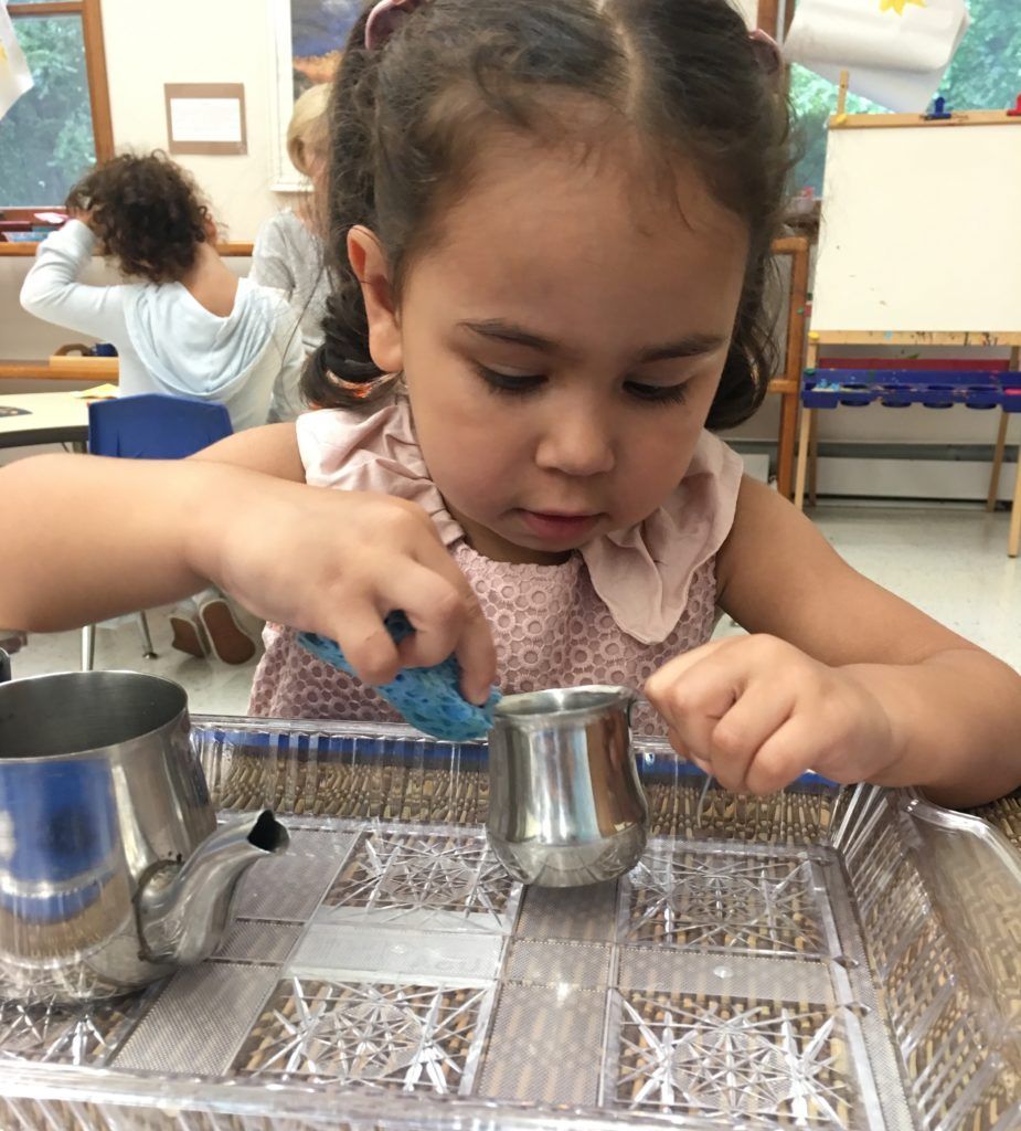 A young girl with braids focuses intently as she carefully pours liquid from a small silver pitcher.