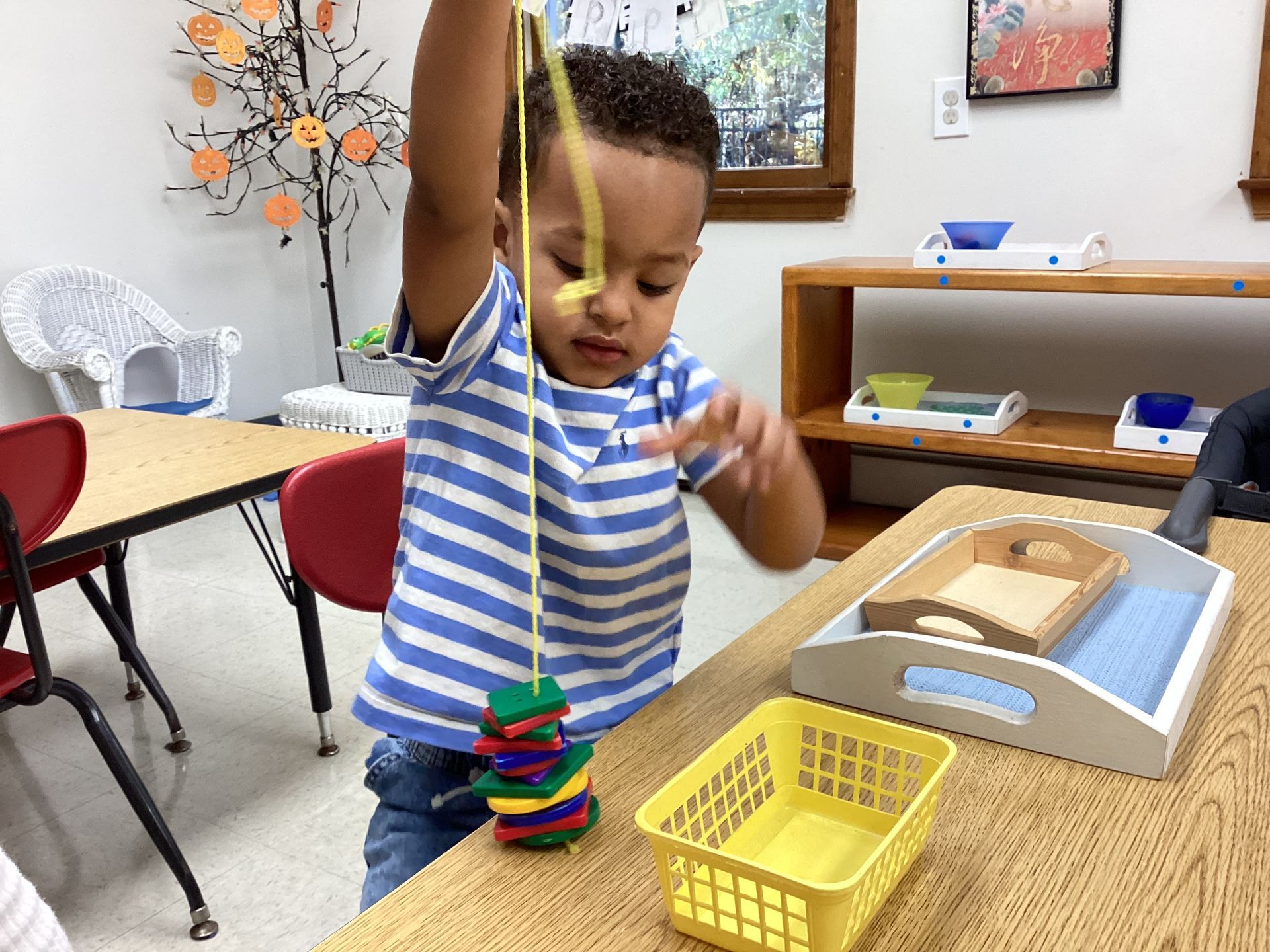 A young child in a blue striped shirt focuses on threading colorful shapes onto a yellow string at a classroom table.