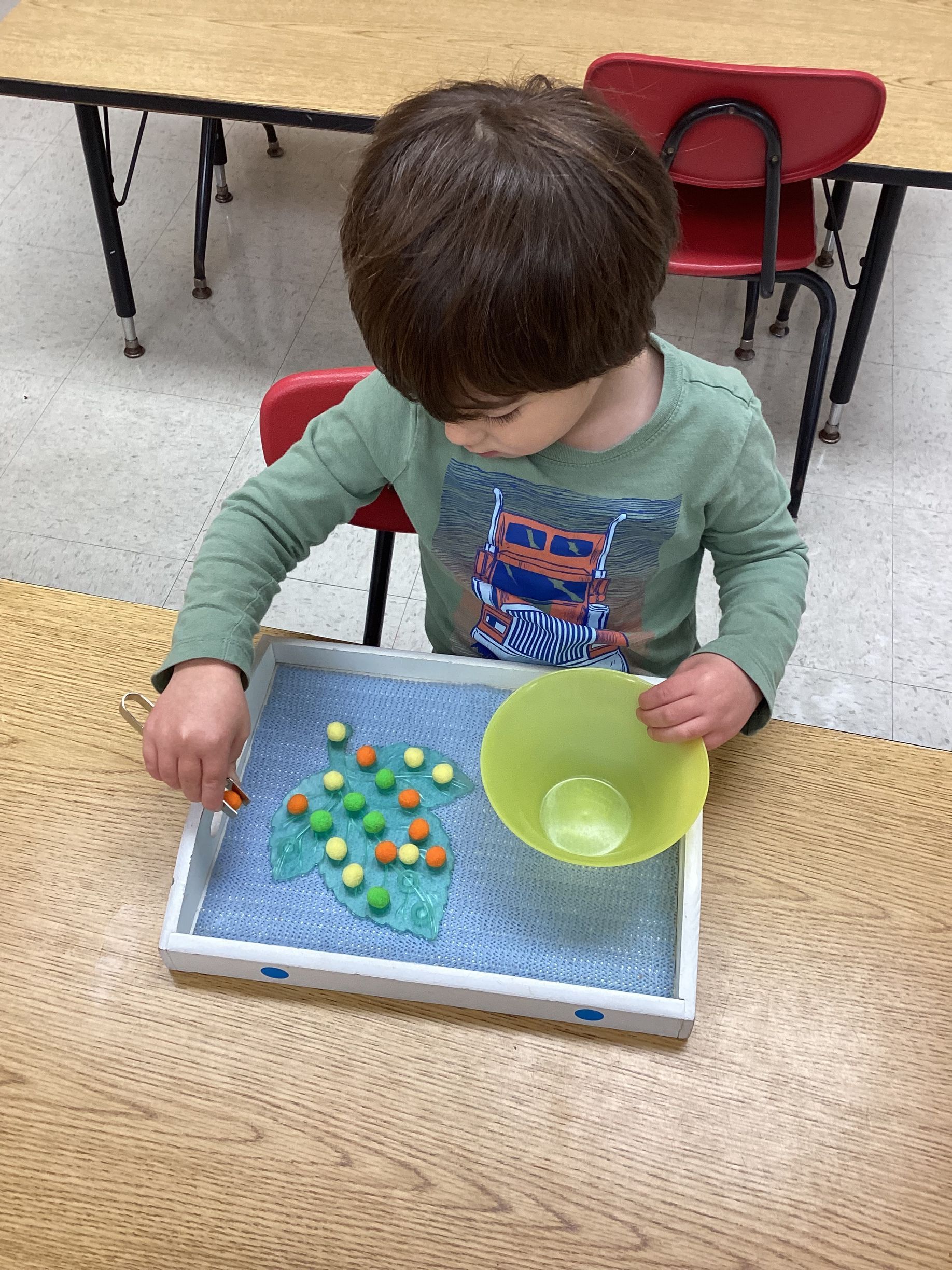 A child wearing a green shirt sits at a table, focused on picking up colorful pom-poms from a textured tray using tweezers.