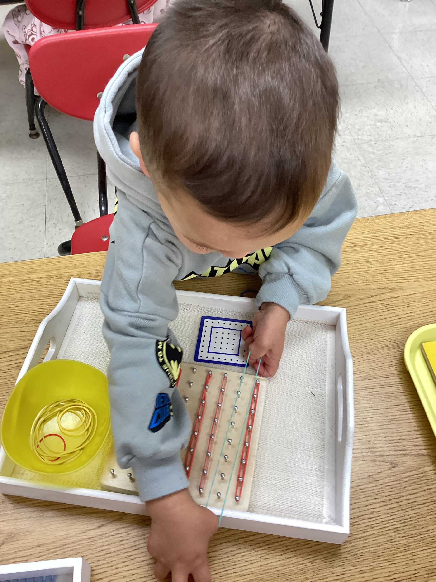 A child in a grey sweater engages with a geometric peg board activity on a wooden table, using rubber bands and pegs.