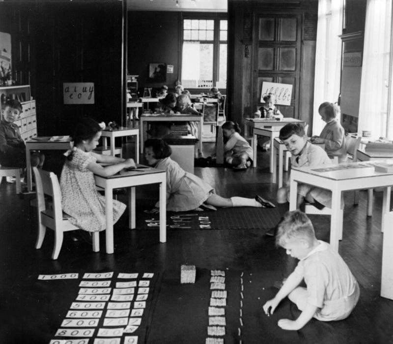 Children in a classroom engage in various activities at small tables, using counting cards on the floor.