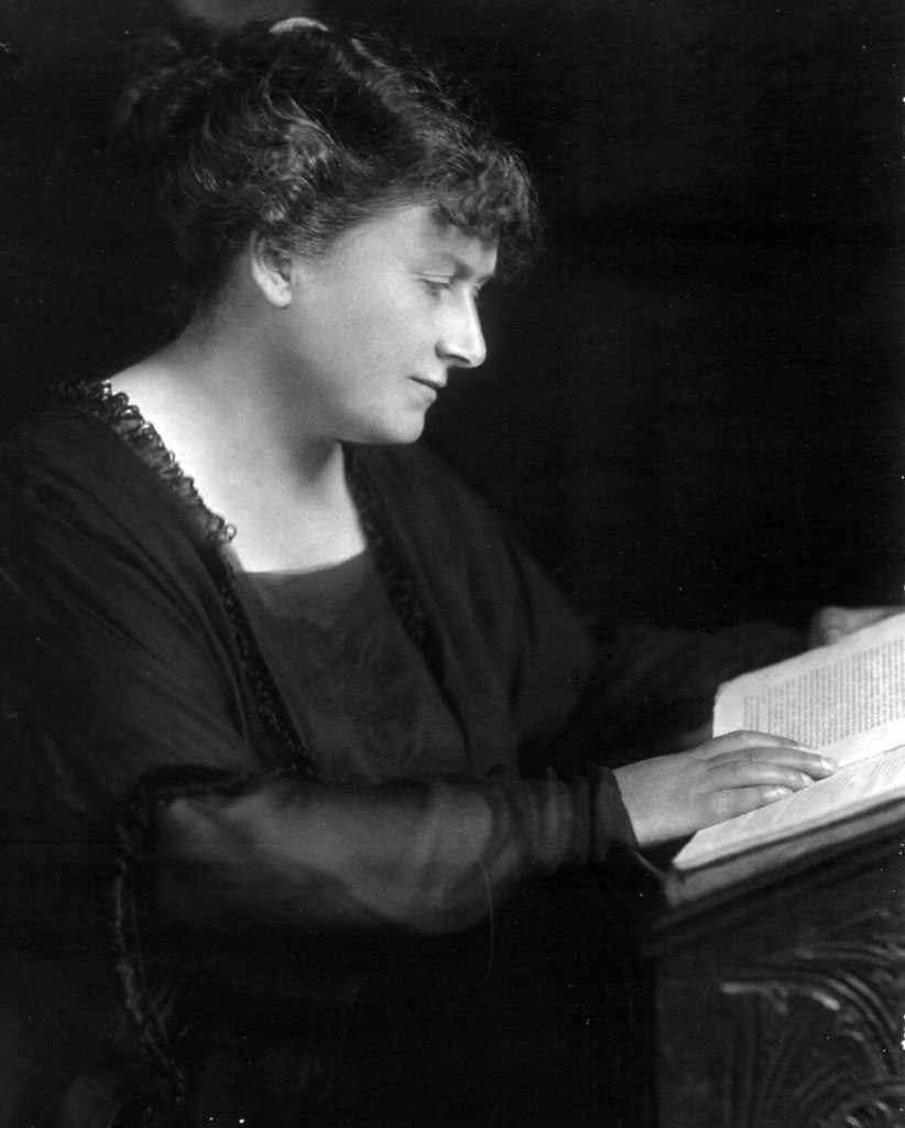A woman with short hair sits at a wooden desk, reading a book, dressed in a dark blouse.