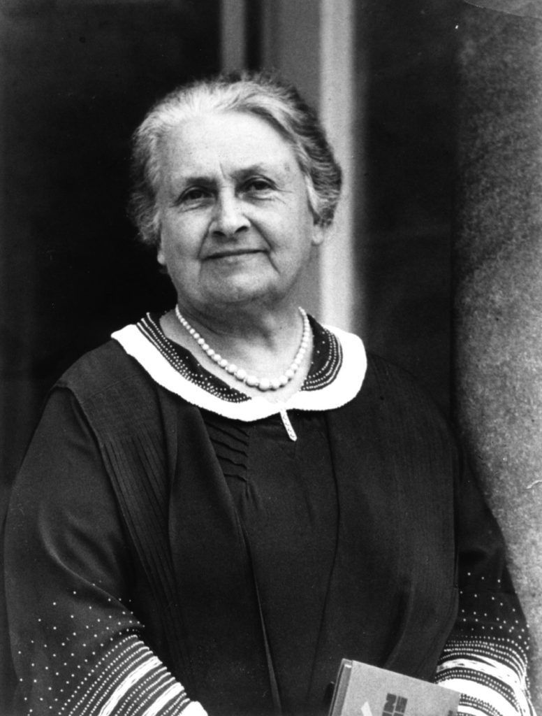 An elderly woman with gray hair stands outside, wearing a dark dress with a white collar and a pearl necklace.