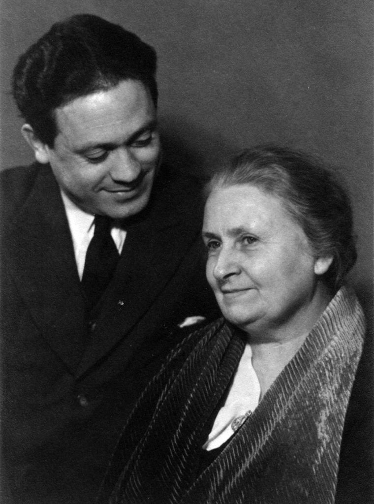 A man, in a suit, gazes warmly at an older woman wrapped in a shawl. She smiles slightly, conveying a sense of warmth.