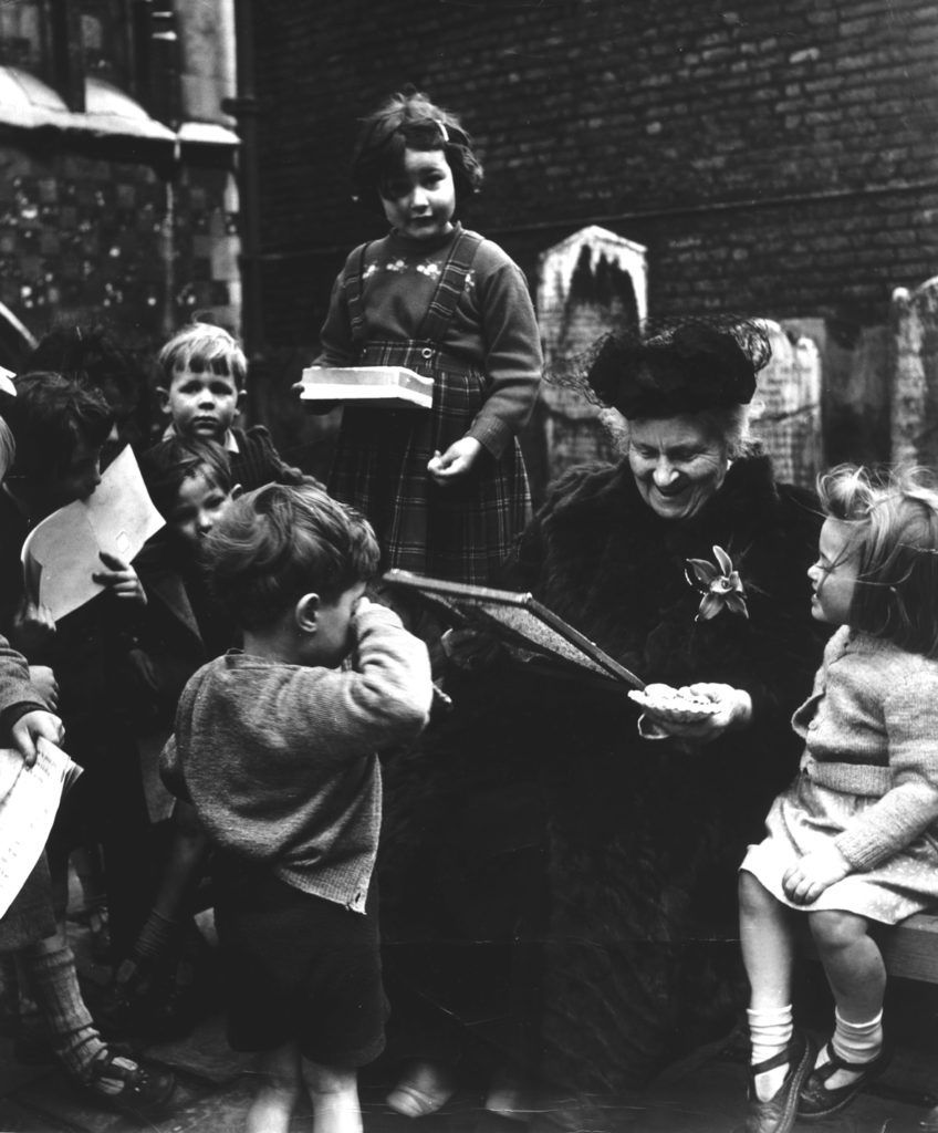 An older woman, dressed warmly, smiles while seated, surrounded by young children in a brick courtyard.