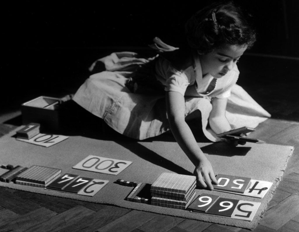 A young girl in a dress engages with Montessori math materials on the floor. She is very focused on her acticity.