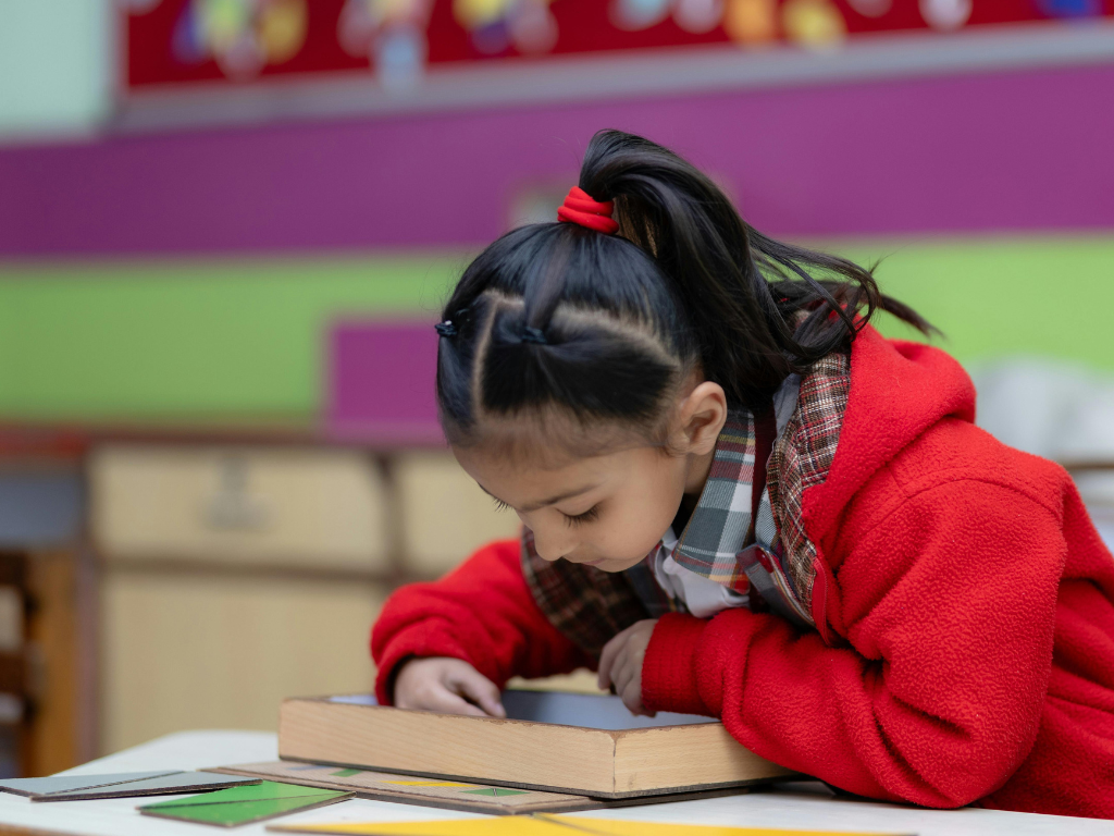 A young girl in a red jacket leans over a desk, focused on a book. The background is a colorful classroom.