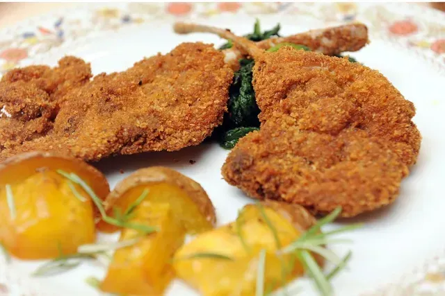 Due costolette d'agnello impanate servite con spicchi di patate arrosto e verdure saltate in padella su un piatto decorato.