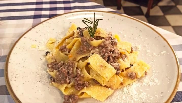 Un piatto di pappardelle al ragù di carne con guarnizione di rosmarino, servito su una tovaglia a quadri blu e bianchi.