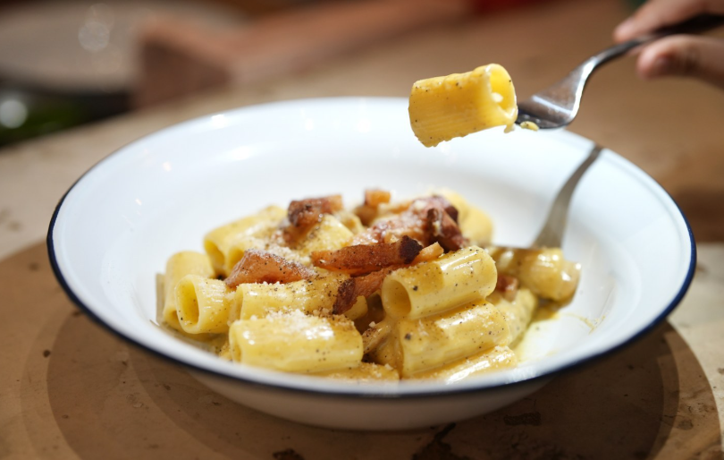 Una forchetta sorregge un rigatone sopra una ciotola bianca piena di pasta cremosa, pezzetti di pancetta e formaggio grattugiato.