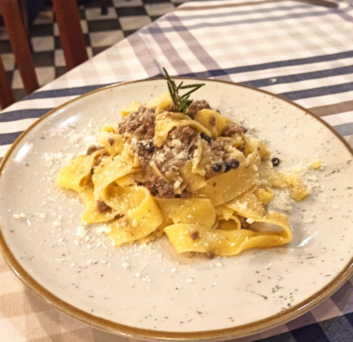 Un piatto di pappardelle al ragù, guarnite con formaggio grattugiato e un rametto di rosmarino, servito su una tovaglia a quadri.
