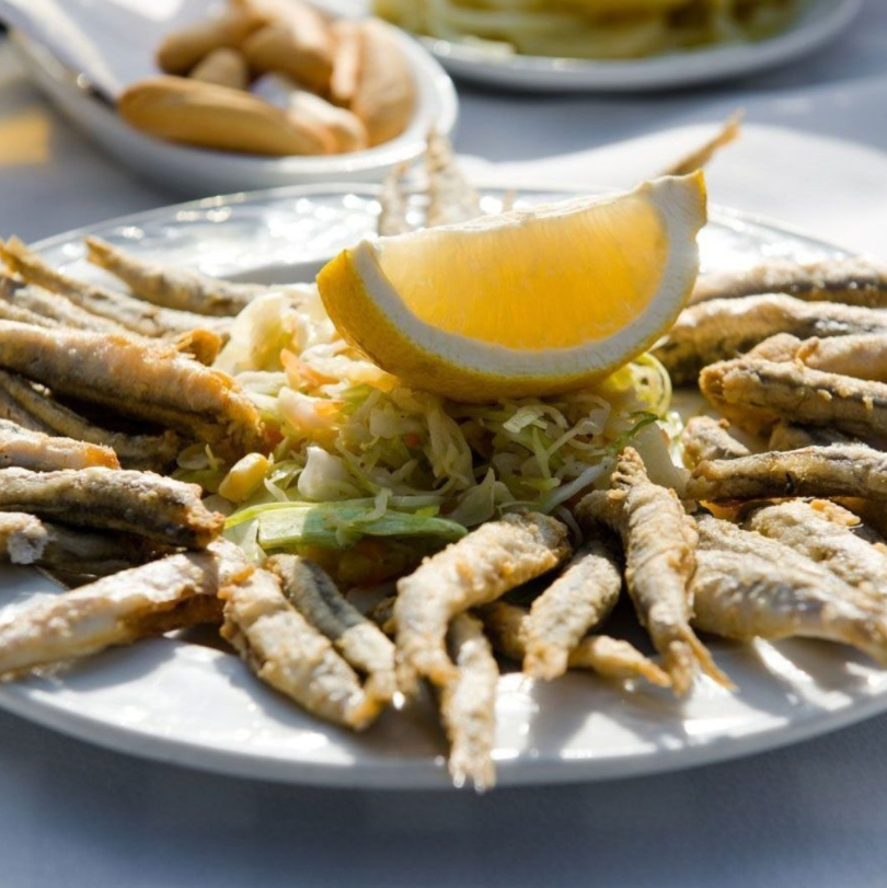 Un piatto di piccoli pesci fritti croccanti, servito con uno spicchio di limone e un contorno di verdure tritate su una tovaglia bianca.