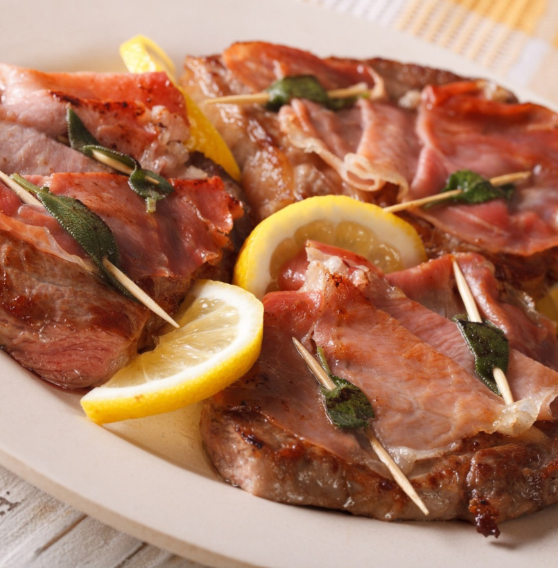 Saltimbocca alla romana con prosciutto crudo a fette sottili, foglie di salvia e spicchi di limone su un piatto.