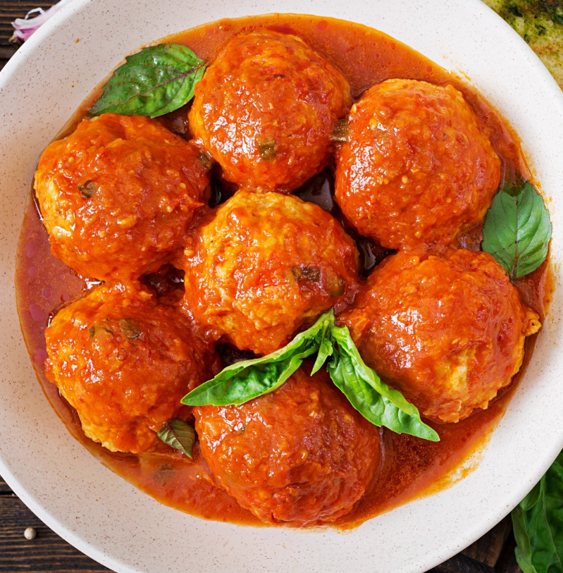 Una ciotola contenente sei polpette cotte, ricoperte da una densa salsa di pomodoro rosso e guarnite con foglie di basilico fresco.