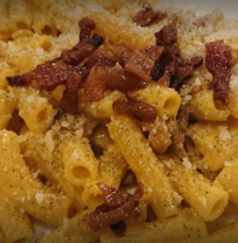 Primo piano di pasta con pancetta croccante, formaggio grattugiato e pepe nero, ricoperta da una cremosa salsa gialla.