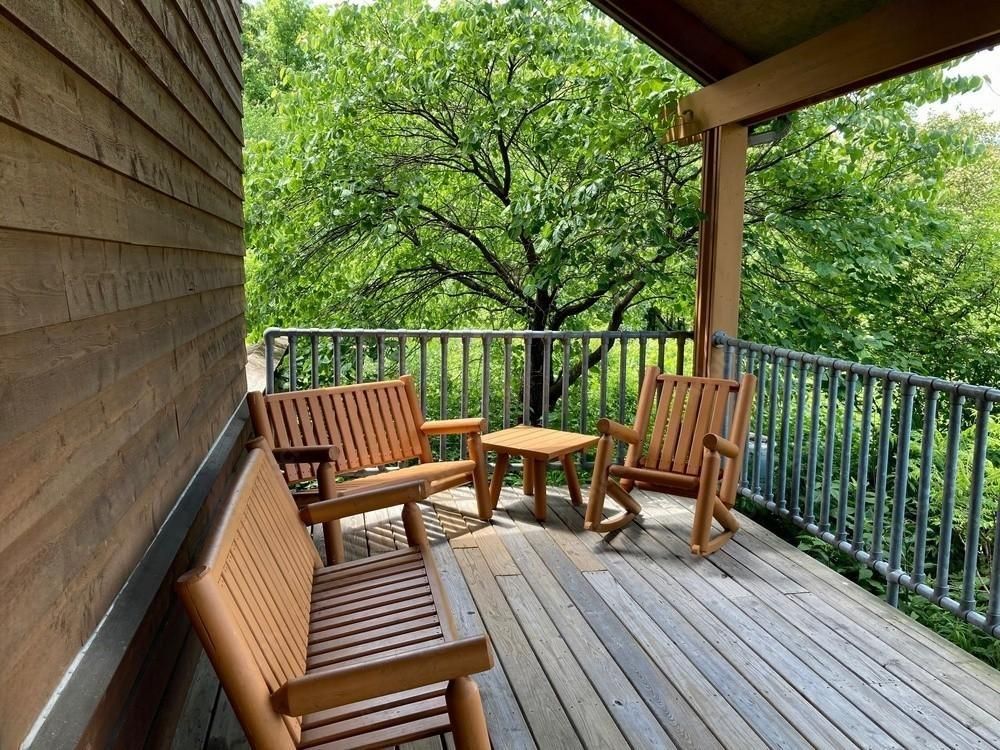 A wooden deck featuring two benches, a small side table, and a rocking chair, overlooking a lush green forest.