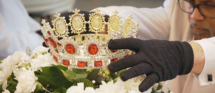 Gloved hands laying an ornate crown atop a spray of flowers and greenery.