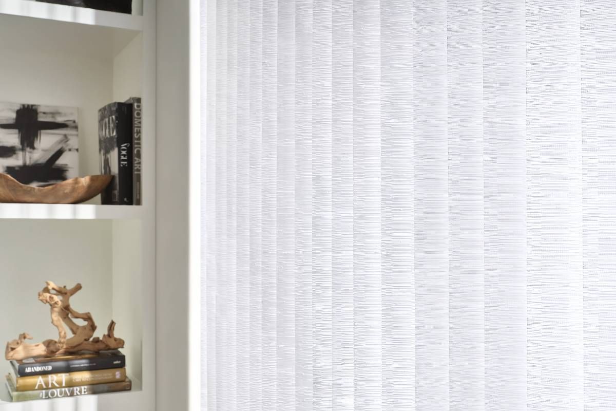 A detailed image of white and textured Hunter Douglas Somner® Vertical Blinds beside a bookcase at Maple Drapery & Carpet