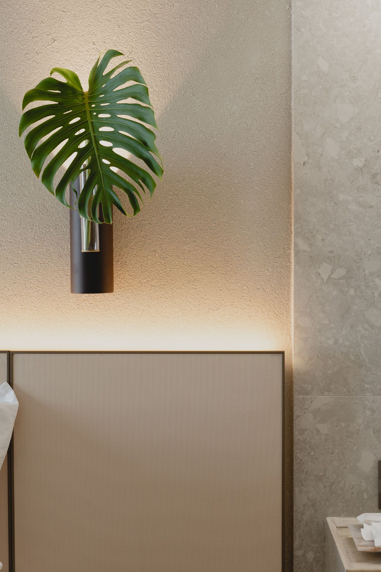 Folha de Monstera em um vaso fixado na parede contra uma parede bege texturizada com iluminação suave acima de uma cabeceira estampada.