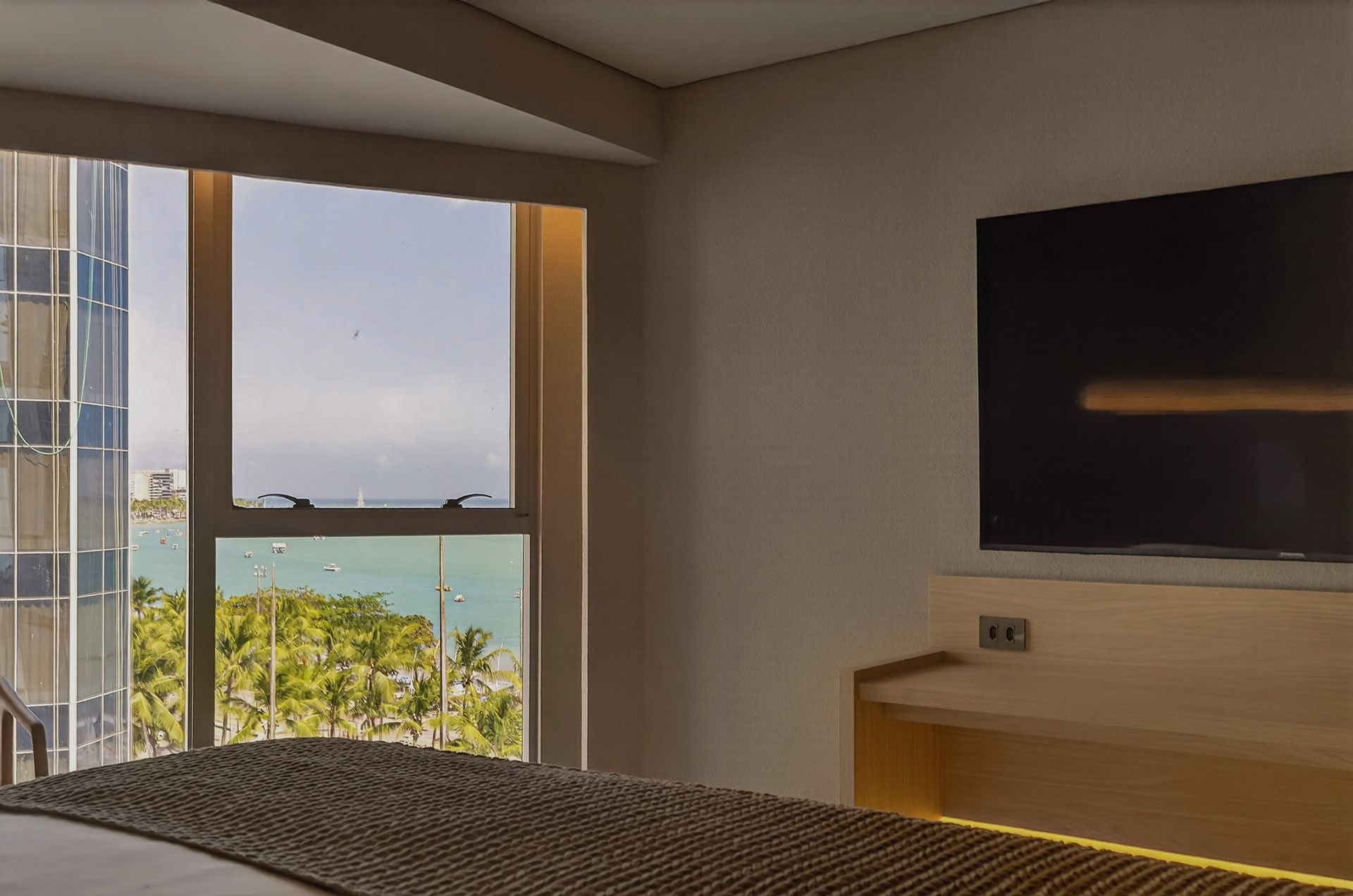 Quarto com vista para o mar a partir da janela, TV grande montada na parede acima da bancada de madeira.