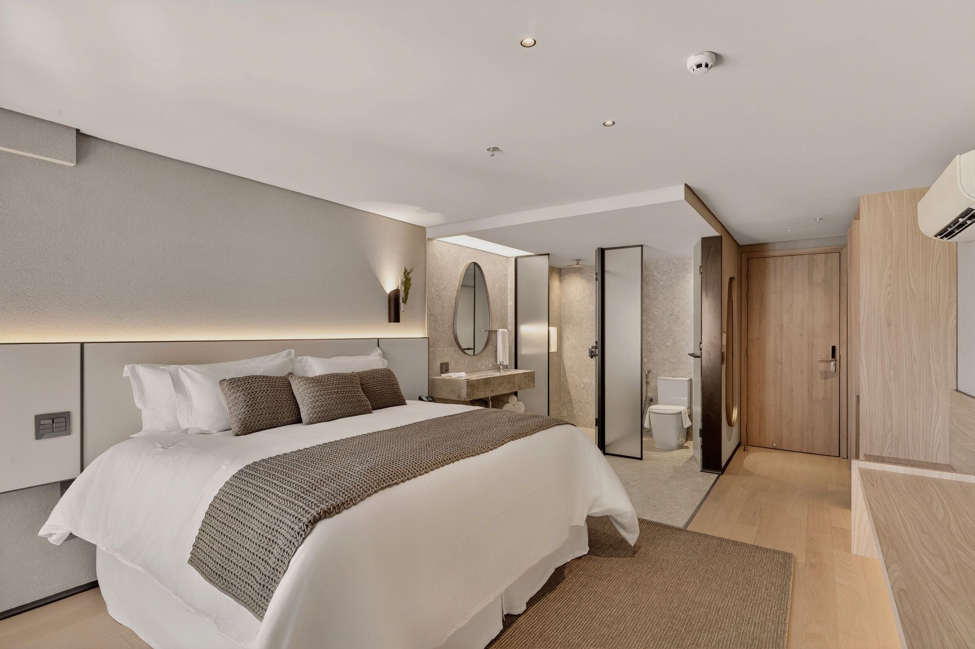 Quarto de hotel moderno com cama, mesa, cadeiras e banheiro. Cores neutras e iluminação natural.