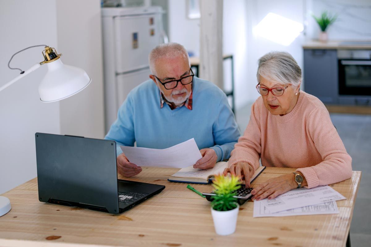 An elderly couple is sitting at a table looking at papers and a laptop.
