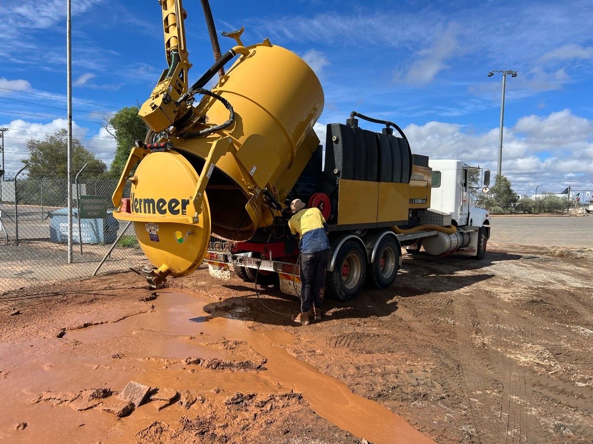 Yellow concrete mixer truck pouring wet concrete at a muddy construction site, worker standing nearby