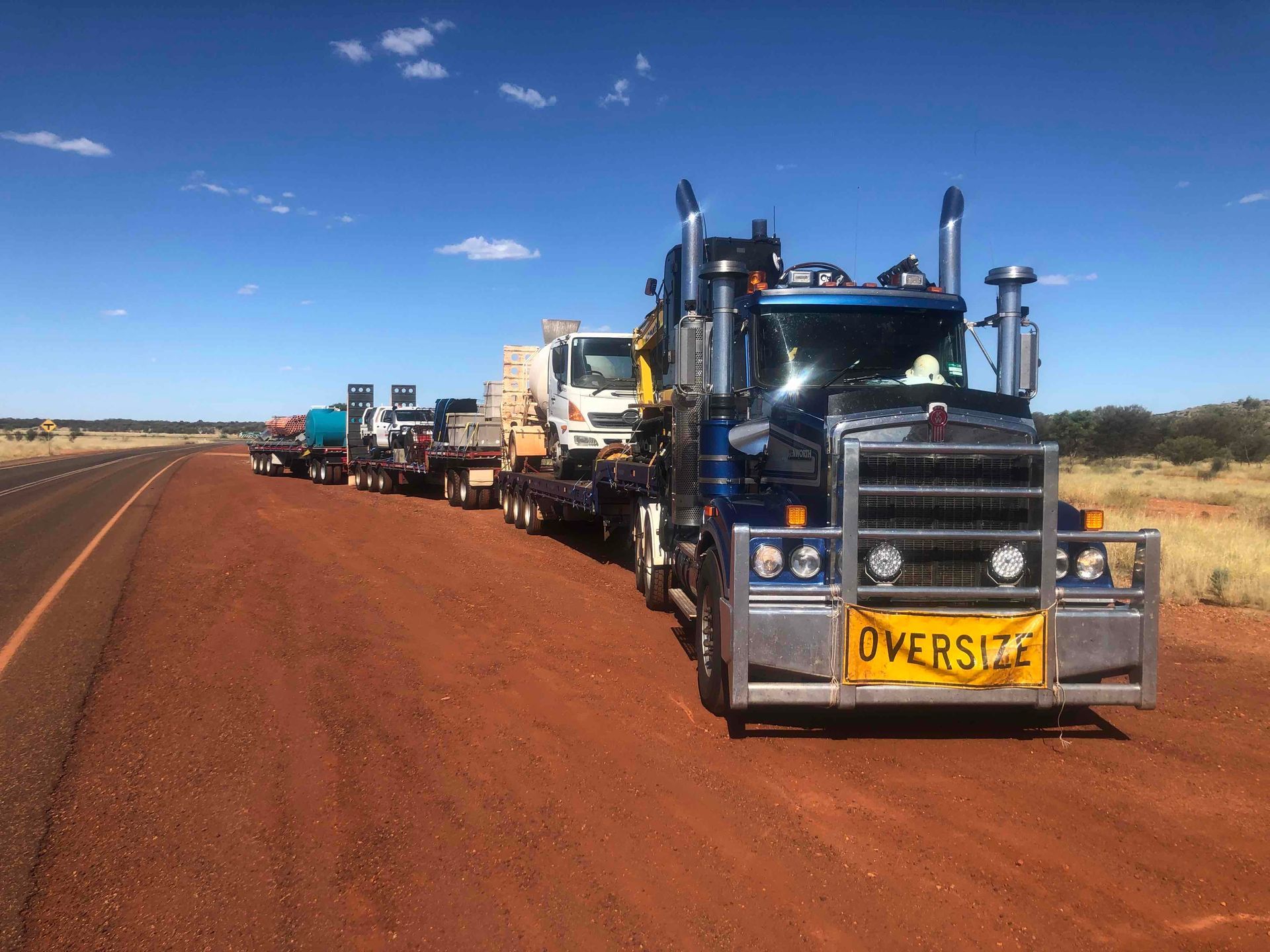 Convoy of heavy trucks on a red dirt road under a blue sky, lead truck marked “OVERSIZE”