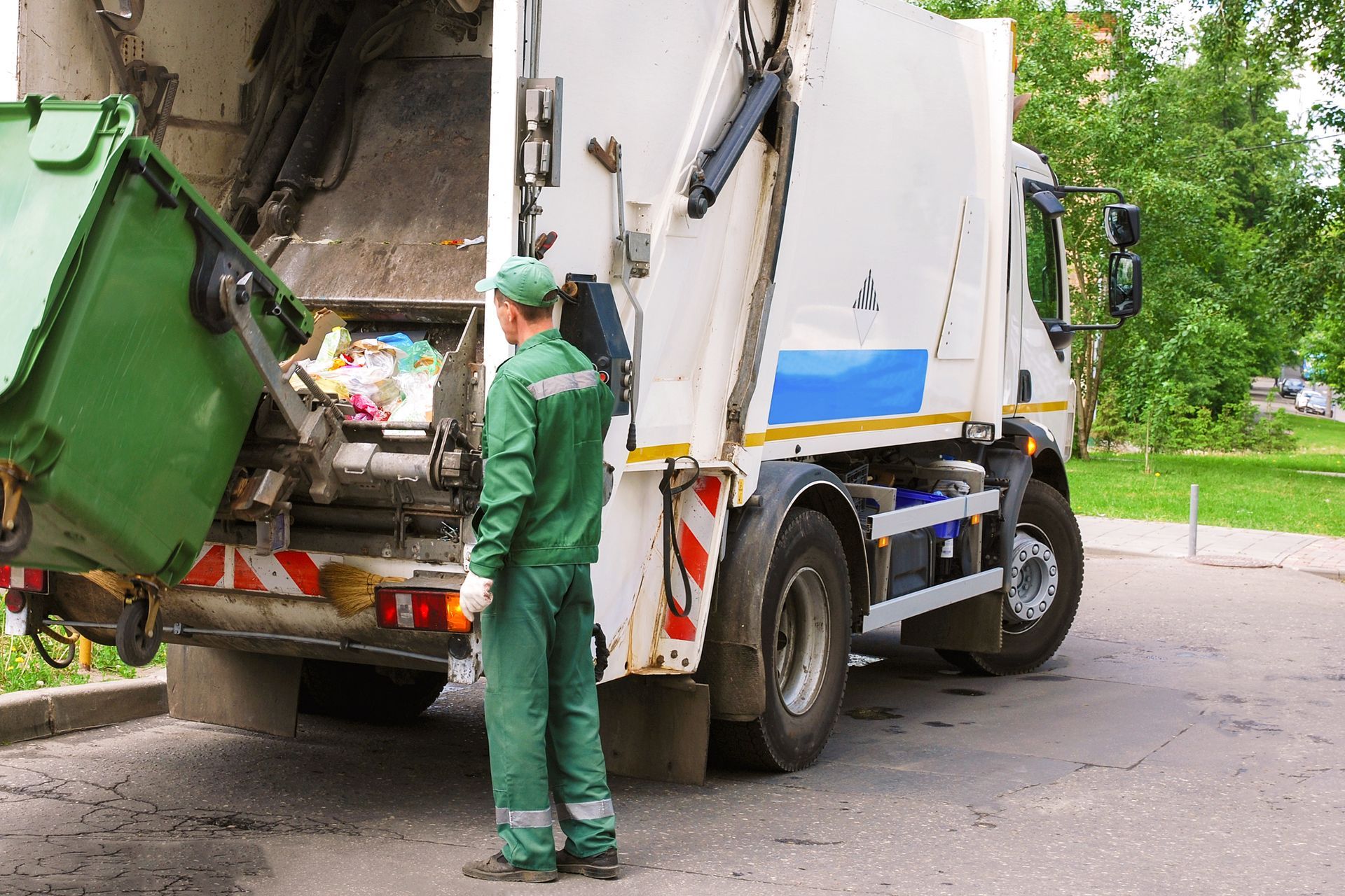 A sanitation worker empties a green trash bin into a garbage truck on a residential street.