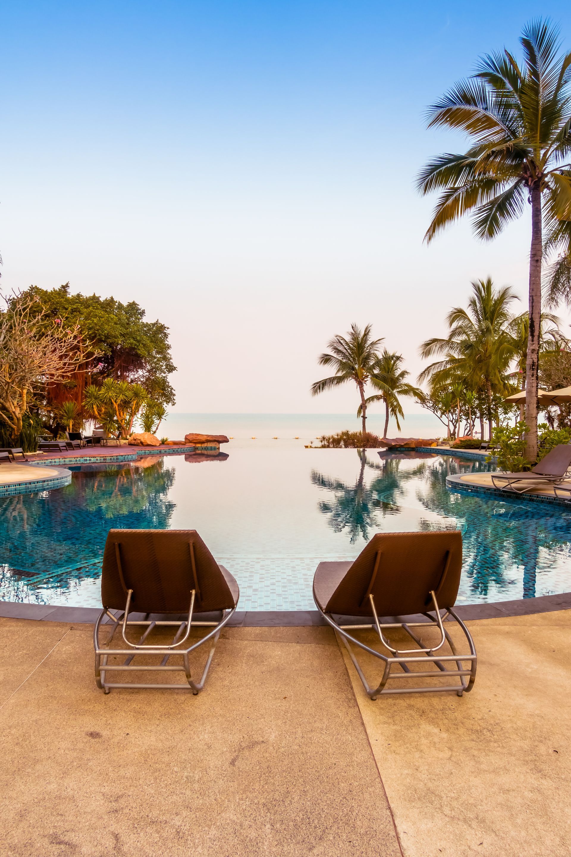 A swimming pool surrounded by chairs and palm trees with a view of the ocean.