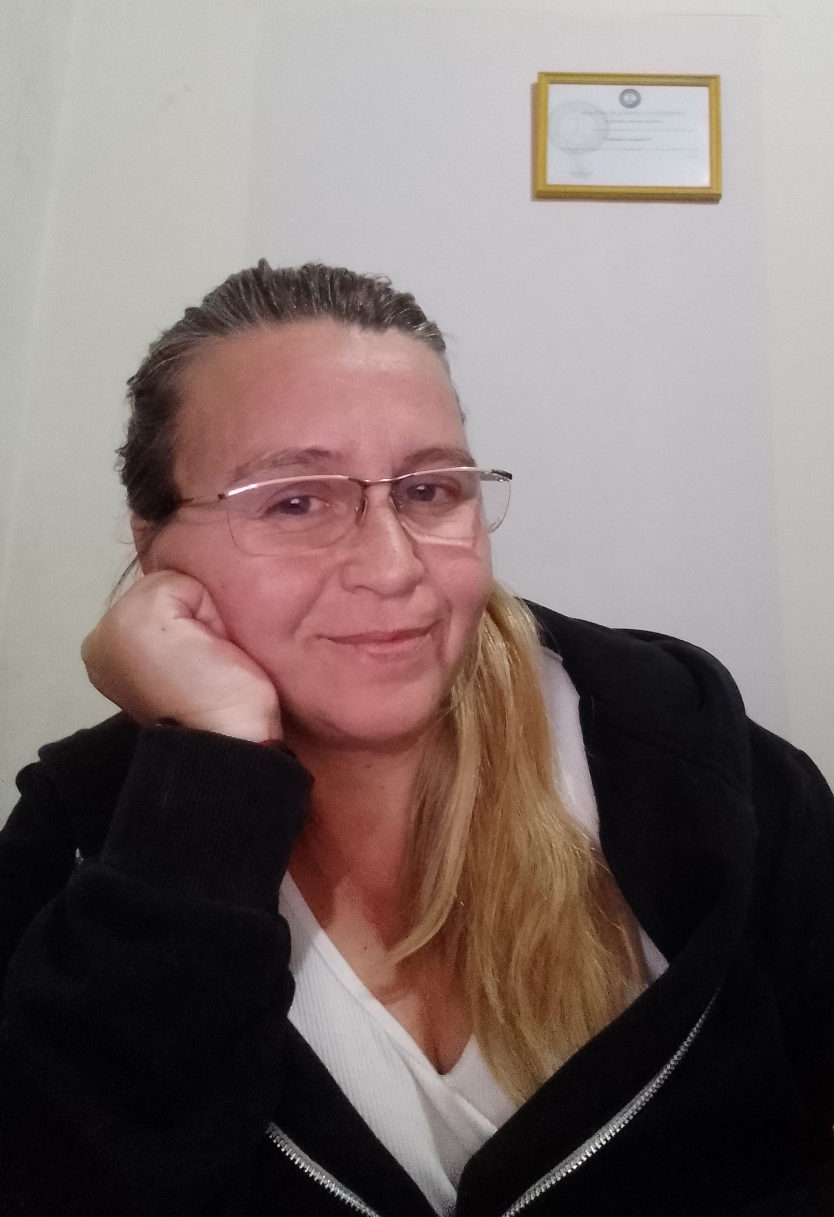 Mujer con gafas, apoyando la cabeza en la mano, sonríe, contra una pared blanca, diploma en la pared.