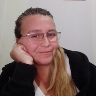 Mujer con gafas, apoya la barbilla en la mano y sonríe. Cabello rubio, con chaqueta negra en el interior.