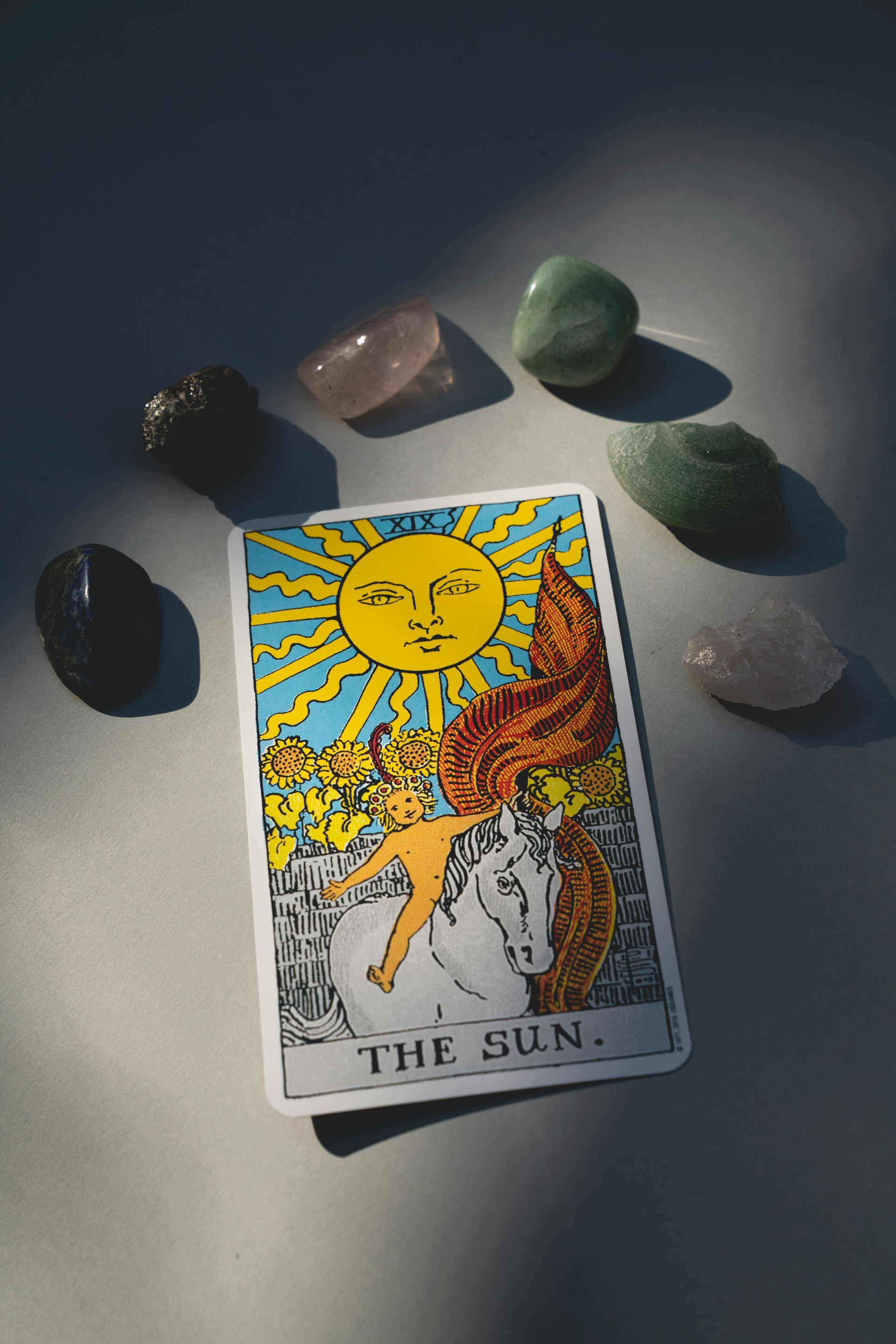 Carta del Tarot "El Sol" rodeada de cristales sobre una superficie gris, bañada por la luz del sol.