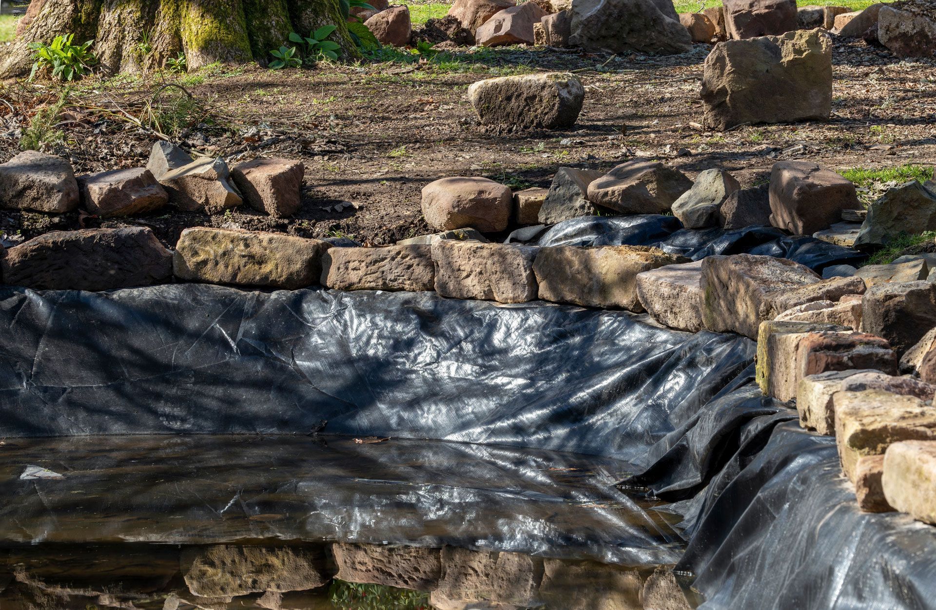 Pond being constructed with black liner and stone edging, surrounded by stone and dirt, under trees.