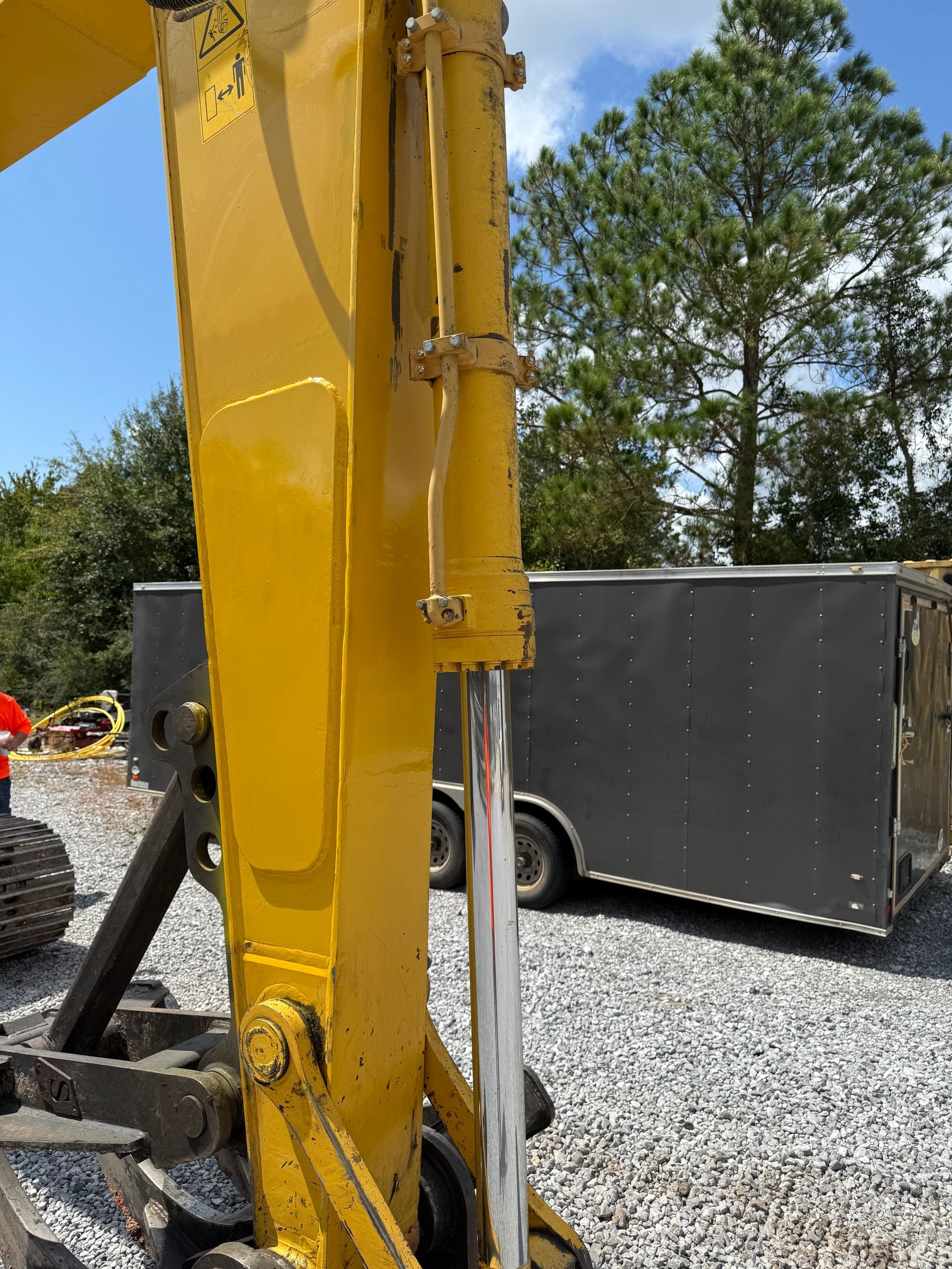 Construction worker on a platform, directing a crane that lifts a large wooden beam. Buildings and cloudy sky in background.