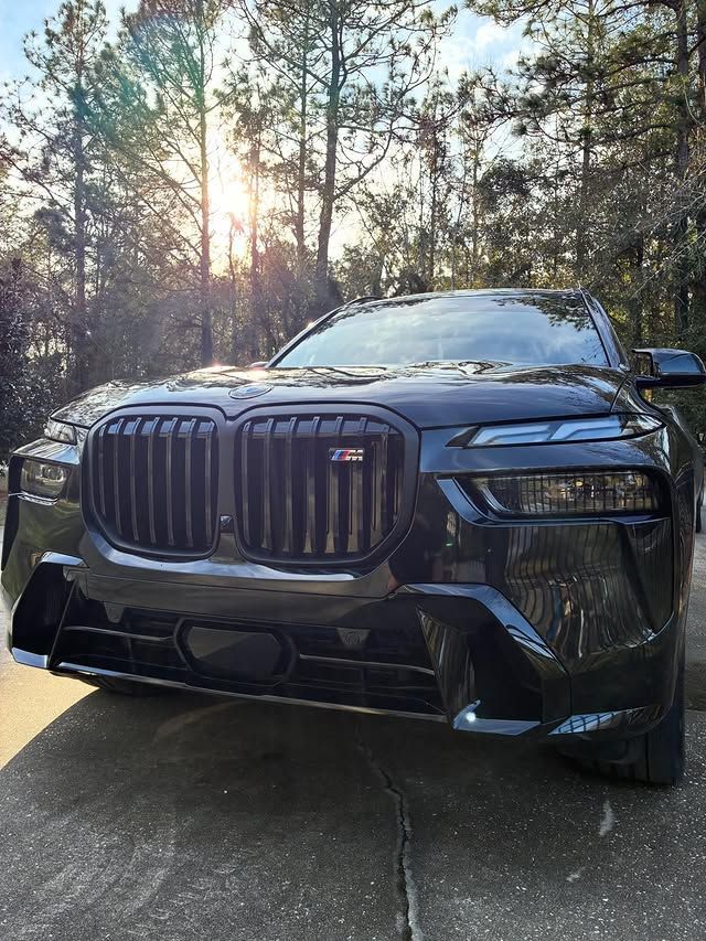 A black BMW SUV front view with a prominent grille and M badge, parked on a driveway with trees in the background.