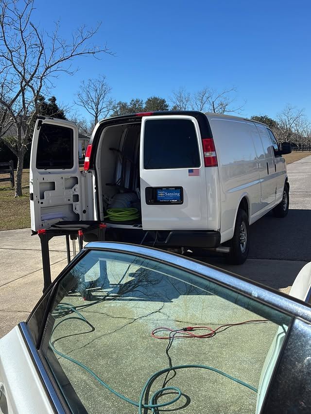 A white work van parked outdoors with doors open, viewed over the foreground of a car windshield.