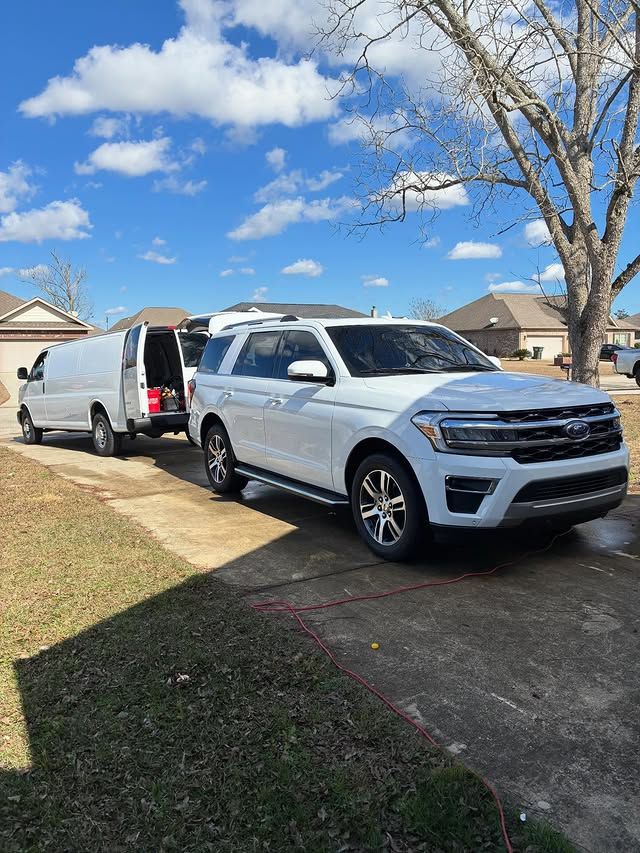 A white SUV is parked in a residential driveway in front of a white work van under a clear blue sky.