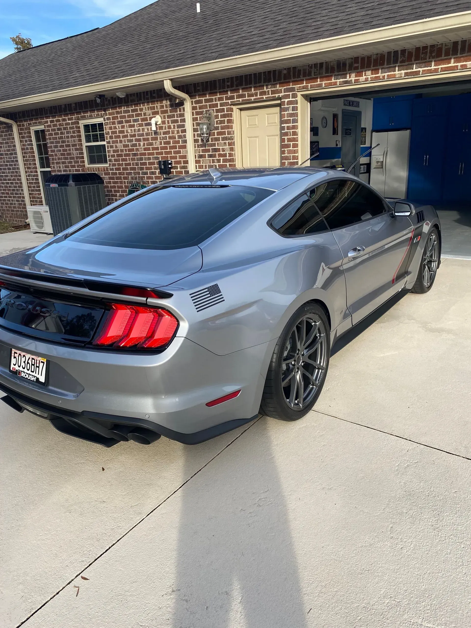 A silver Ford Mustang parked in a residential driveway, featuring a small American flag decal on the rear quarter panel.