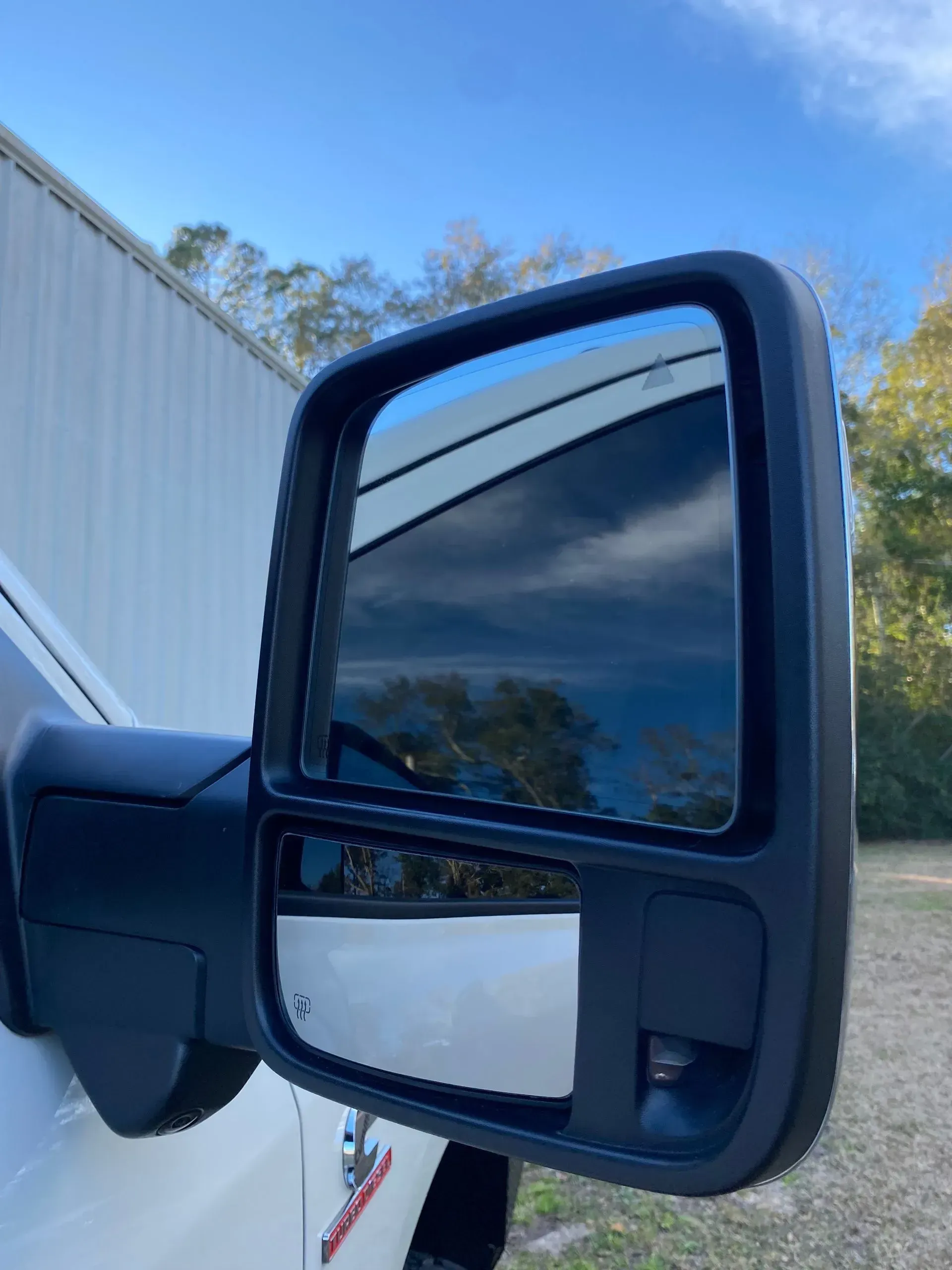 A close-up of a black side-view mirror on a white vehicle, reflecting the sky and surrounding trees.