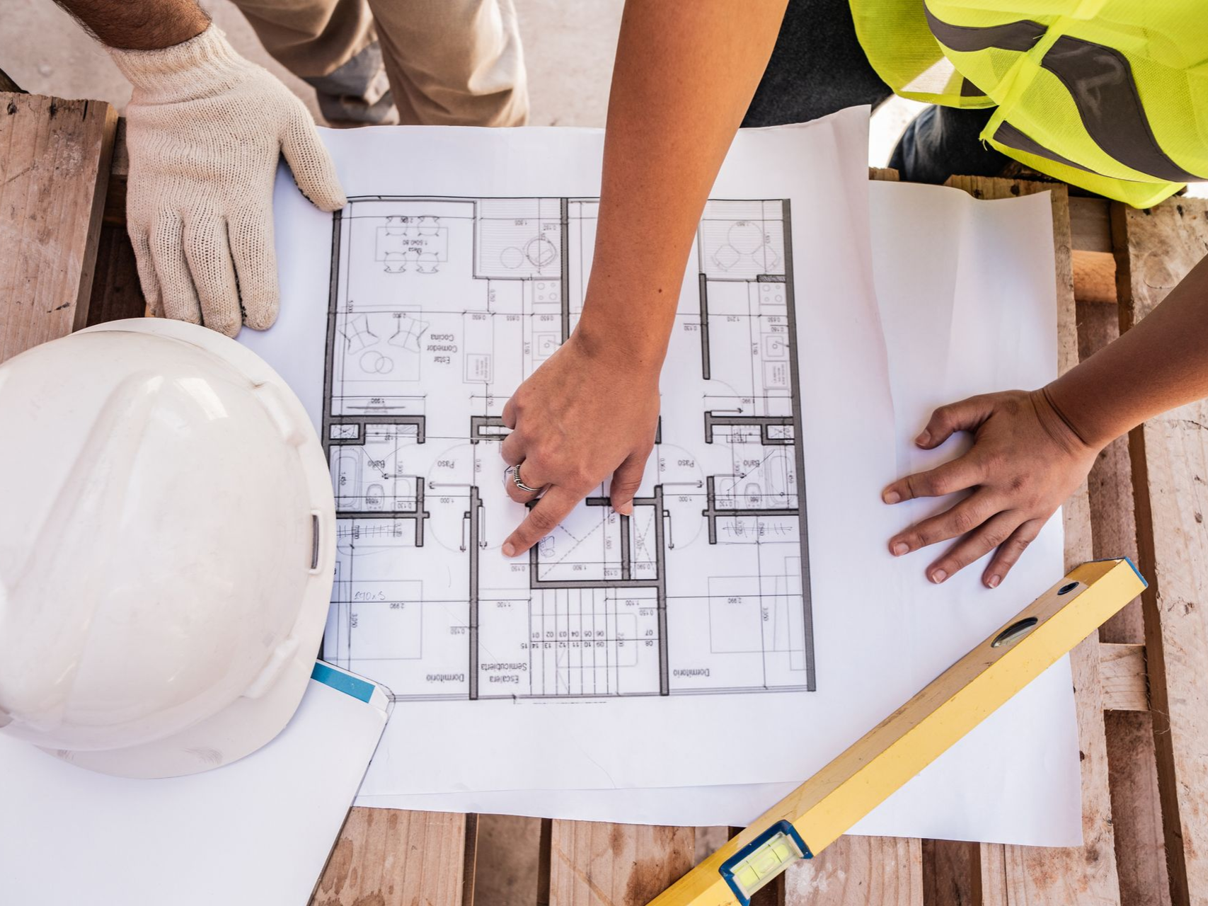 Hands pointing at architectural blueprints on a wooden surface, with a hard hat and level nearby.